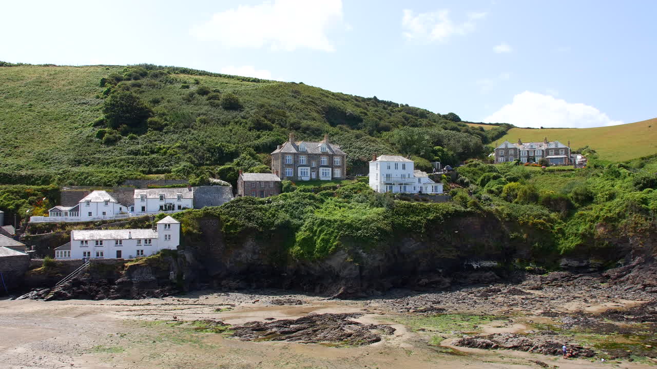 Houses perched along a cliff face overlook a rocky beach, backed by steep green hills and surrounded by dense vegetation and winding footpaths Port Isaac, Cornwall, England