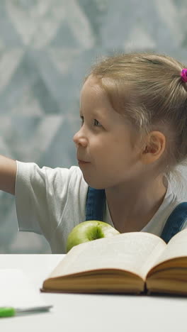 slow motion: adorable little girl with plaits eats green apple and shows thumb-up sitting at white table with open book in spacious light room