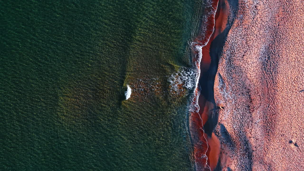 The sun sets over a beach where waves break gently on the shore. Sand meets water creating patterns along the coastline. The scene captures the beauty of nature