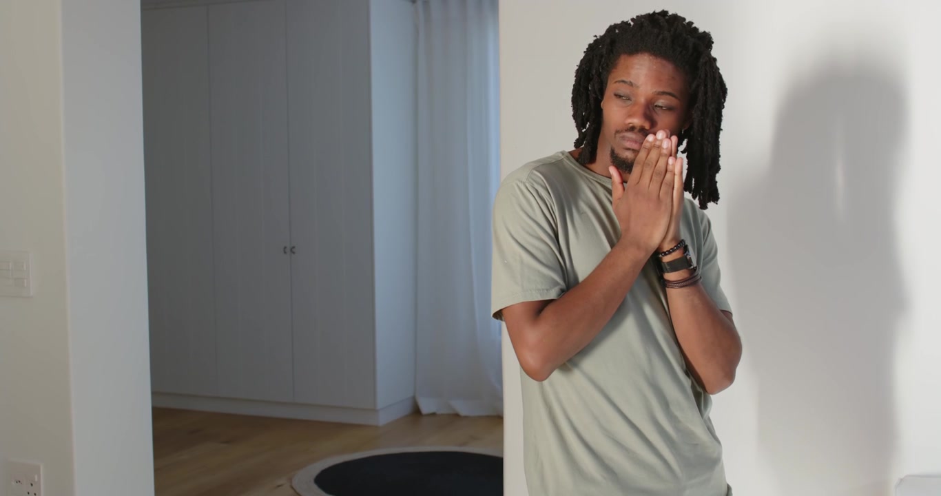 African American man shielding from light, pressing palms, peeking, showing watch by white wall
