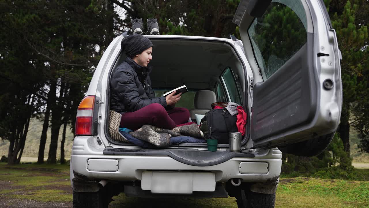 Campeggiatore felice che si gode la natura leggendo un libro nel bagagliaio di un SUV