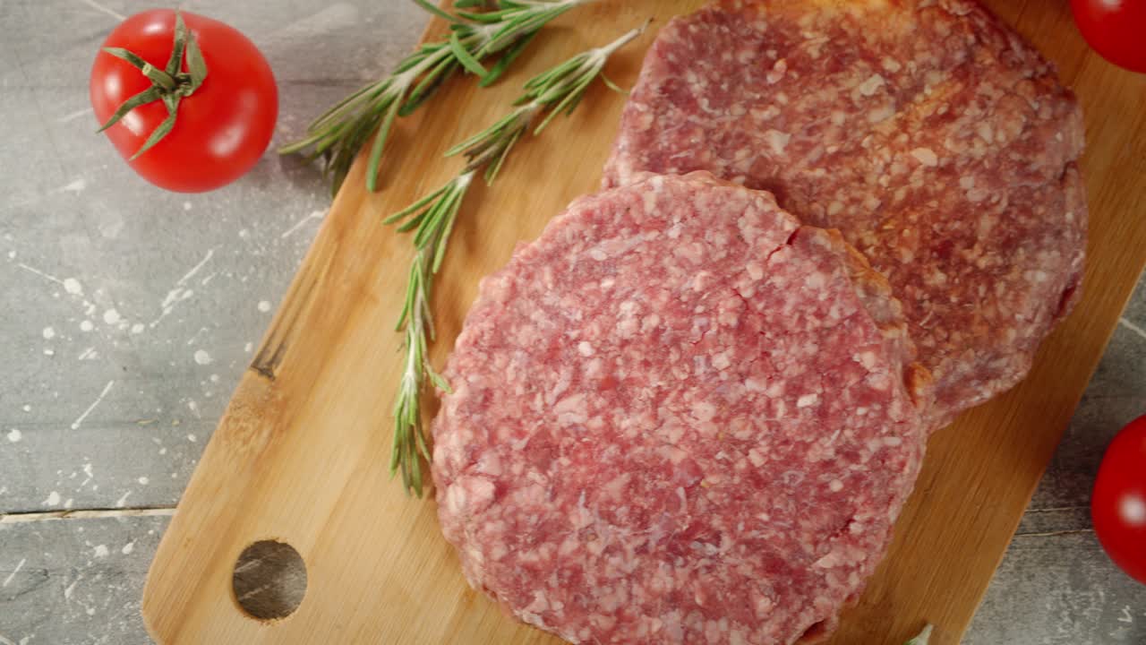 Raw burger with rosemary and tomatoes on cutting board.