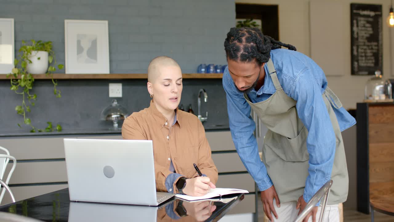 Diverse coworkers working in cafe with one writing notes while other approaching reviewing them