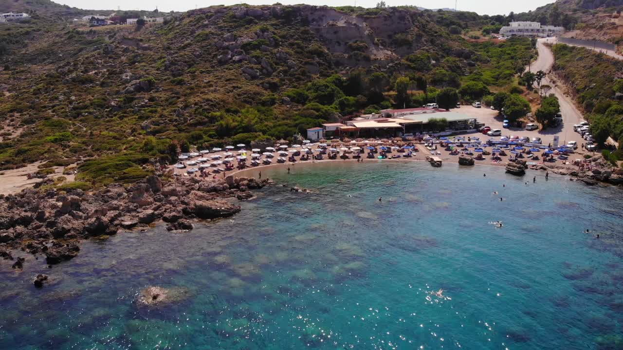 wide aerial view of Anthony Quinn Bay with its azure blue waters and a great beach full of umbrellas and sun beds with the bars and restaurants in the background