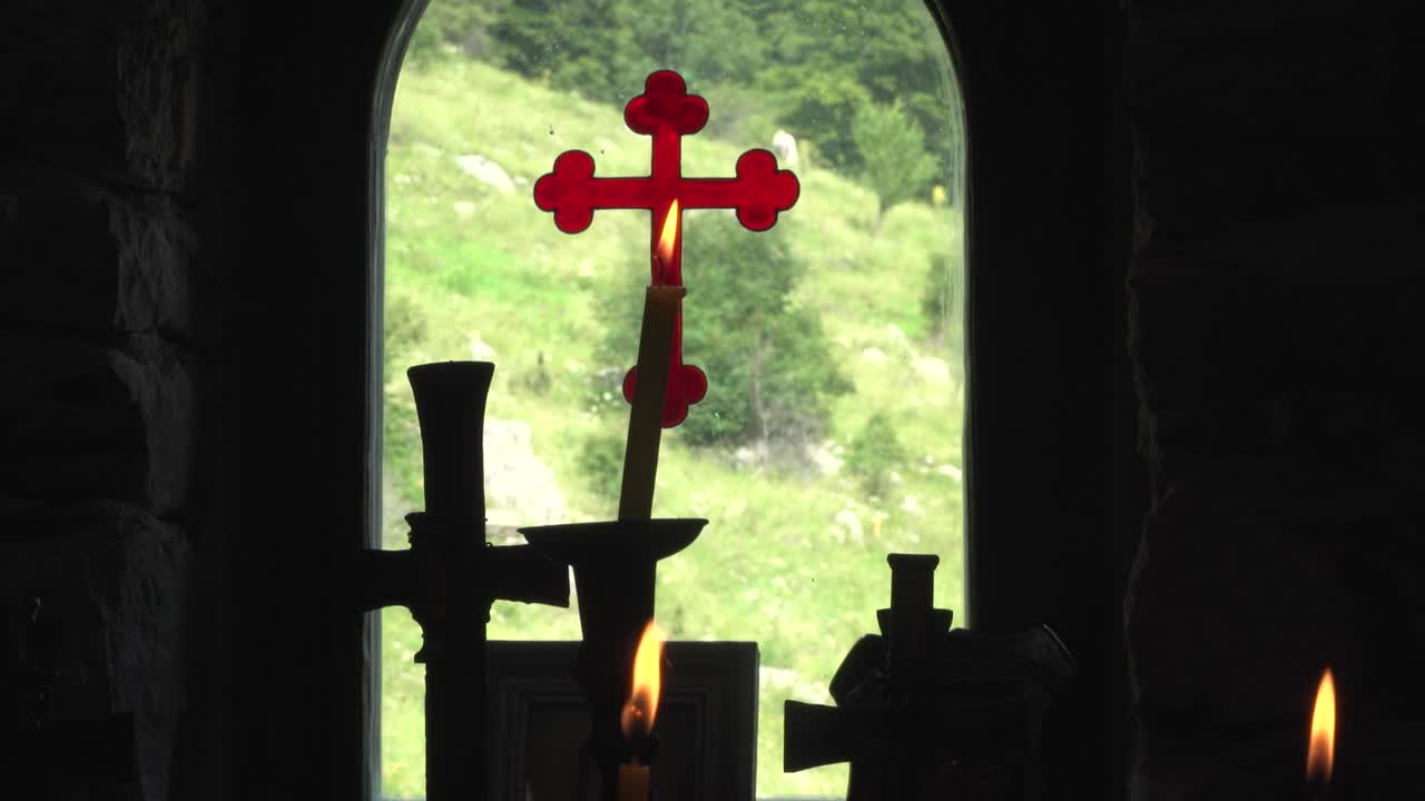 View from inside out through the window of a Bulgarian Orthodox Chapel. Burning candles against a background of a red cross painted on glass. Eastern Orthodox Chapel high in the mountains. Silhouette.