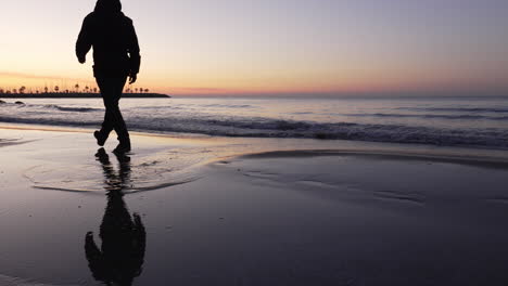 Silhouette-of-man-walking-on-beach-during-sunset,-jumping-over-puddle