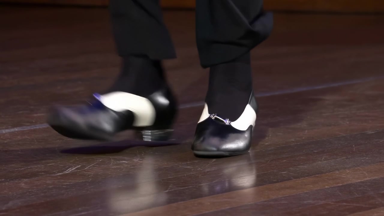 Primo piano di scarpe da tip tap su palco di legno, riflessi sul pavimento lucido, pantaloni neri, colpi ritmici di tacco e punta