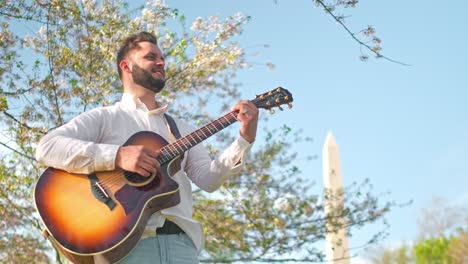 Adult-man-and-musician,-singer,-guitarist,-and-artist-is-standing-at-a-park-singing-a-song-and-strumming-his-guitar-with-the-washington-monument-in-the-background