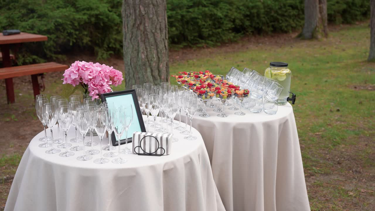 Barra de bebidas para fiesta al aire libre con copas de champán, fruta fresca en vasos de cóctel, dispensador de agua con limón y decoración floral, dispuestas sobre mesas redondas cubiertas de blanco