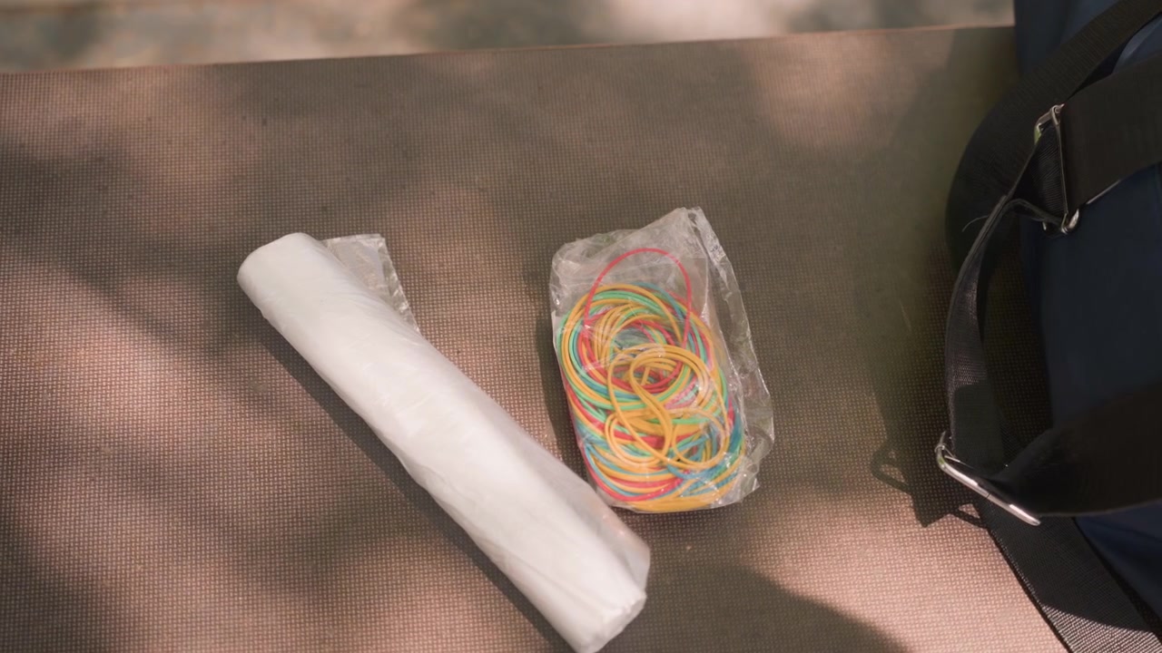 Close up of woman hands placing colorful rubber bands and white fabric on outdoor mat next to blue bag. scene shows careful arrangement of crafting materials in soft natural light with fine