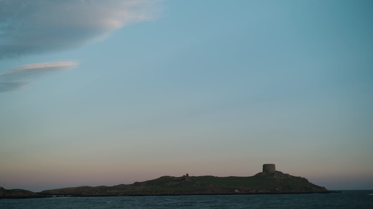 A locked-off shot of Dalkey Island in Dublin, Ireland, with its historic Martello Tower. The sea ripples under a pastel sunset sky, creating a peaceful coastal scene.