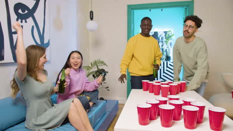 happy multiethnic young friends playing beer pong at home