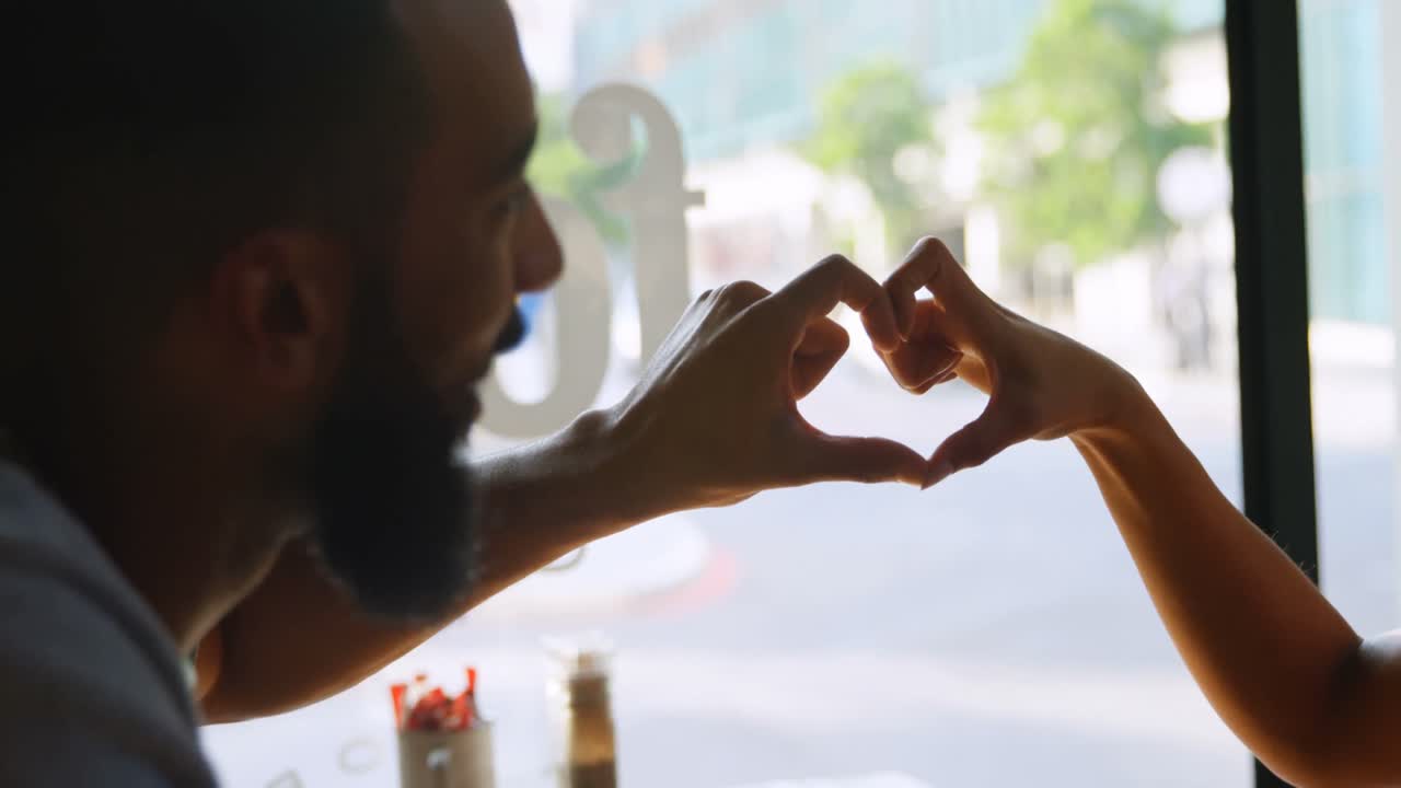 Side view of young mixed-race couple forming heart shape with hands in cafe 4k