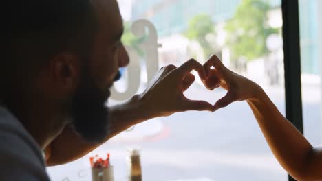 Side-view-of-young-mixed-race-couple-forming-heart-shape-with-hands-in-cafe-4k