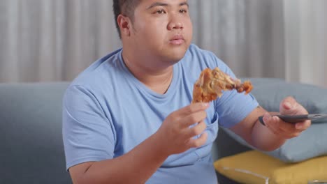 close up of a fat asian man eating fast food having fried chicken while watching tv on a sofa in the living room at home