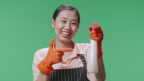 녹색 스크린 스튜디오 에 서 있는 동안 스프레이 를 가리키는 앞옷 을 입은 미소 짓는 아시아 여성 집주인 의 근접 사진