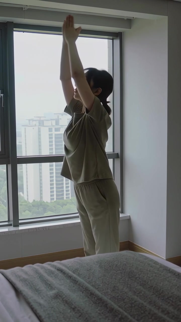 Vertical video: Waking woman in green top and pants raising arms, hinging toward bedroom window