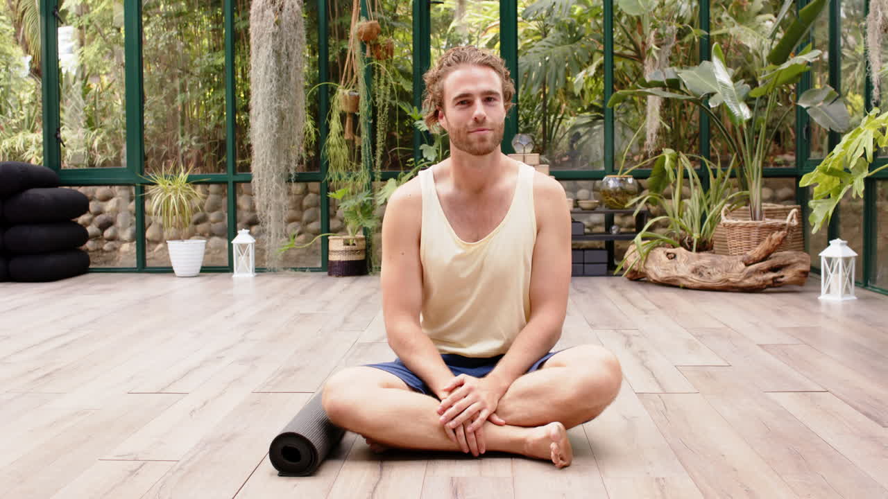 Sitting on floor, man smiling with yoga mat in glass house yoga studio