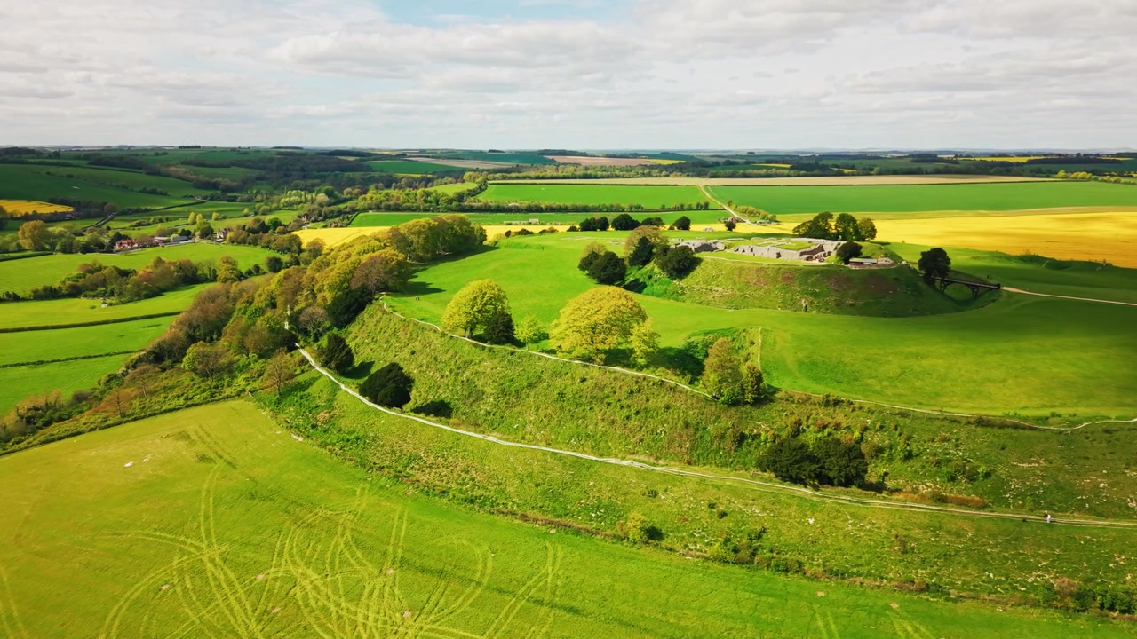 Wide aerial drone shot of Old Sarum near Salisbury in Wiltshire, showing the ancient hillfort earthworks surrounded by green fields, farmland, rural countryside and dramatic cloudy skies.