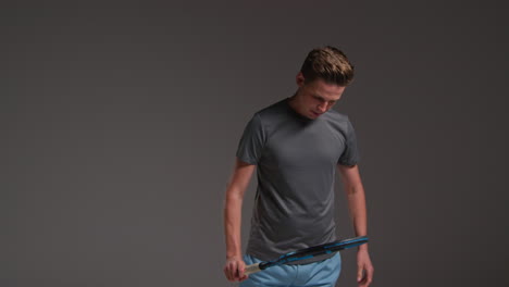 studio portrait of male tennis player holding racket and bouncing ball shot against grey background