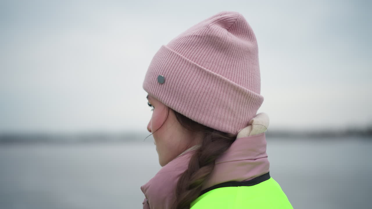 Young woman in pink winter jacket and beanie wearing reflective safety vest speaking outdoors near water on cold overcast day, hair in braid, looking focused and engaged during conversation