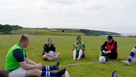 male and female soccer players with coach huddling and sitting in circle on pitch, copy space