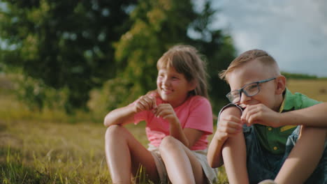 brother and sister sitting together outdoors in warm sunlight, sharing a joyful moment, the boy in glasses looks at his hand curiously while his sister, dressed in pink, smiles behind him