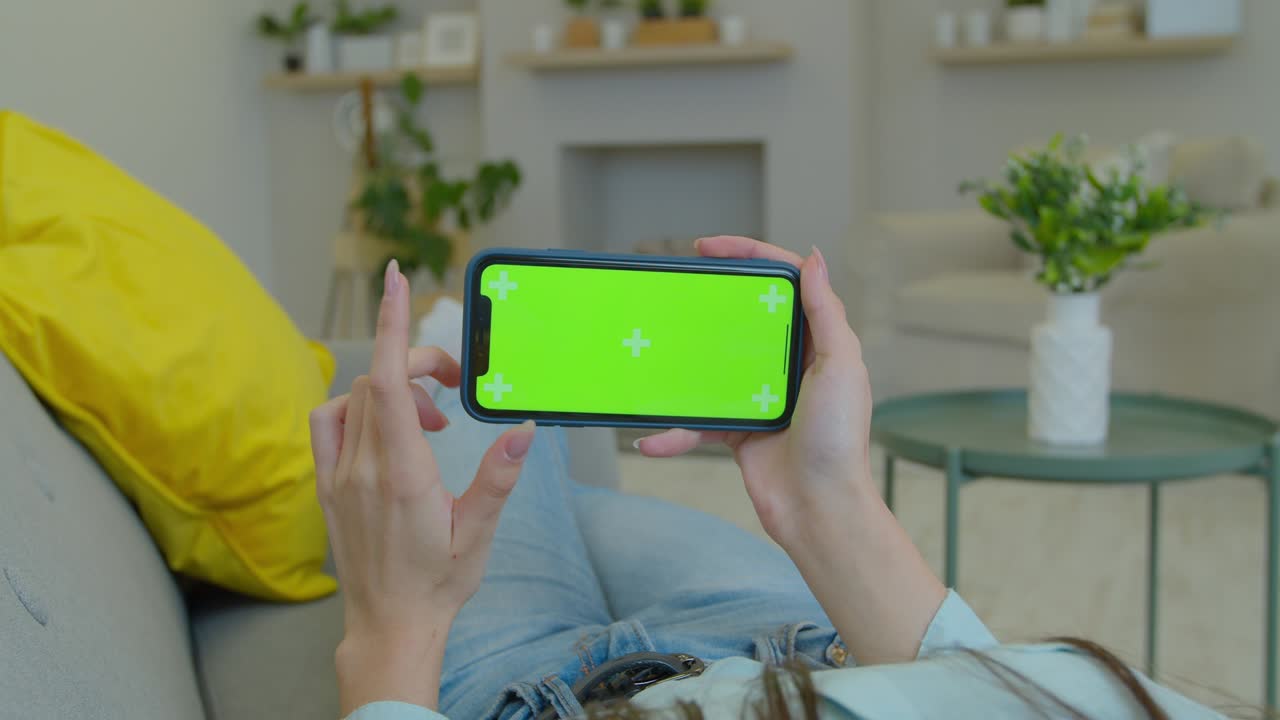 여자친구는 거실에서 녹색 화면으로 수평 풍경 모드에서 스마트폰을 사용하여 영화, 비디오 콘텐츠를 시청합니다. 마커를 추적합니다. 클로즈업.