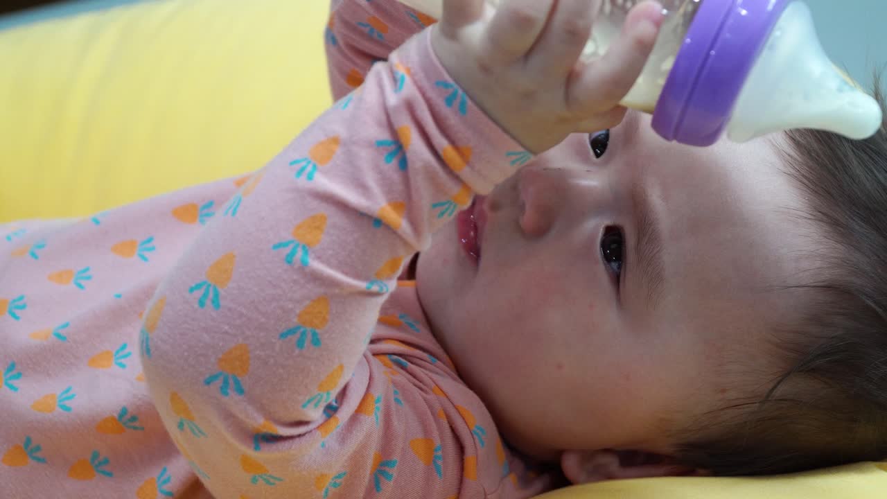 Bambina che tiene e beve latte da un biberon di vetro mentre è sdraiata su un cuscino giallo - primo piano