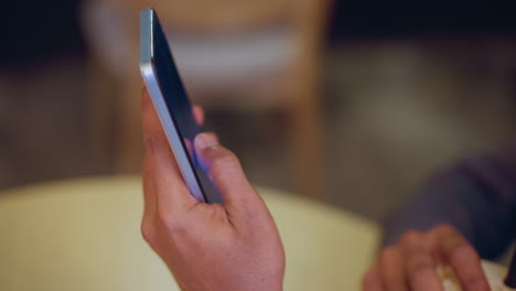 close-up of person holding smartphone with visible lock screen interface while sitting at table, other hand resting near beverage, casual digital moment captured indoors in relaxed atmosphere with soft lighting