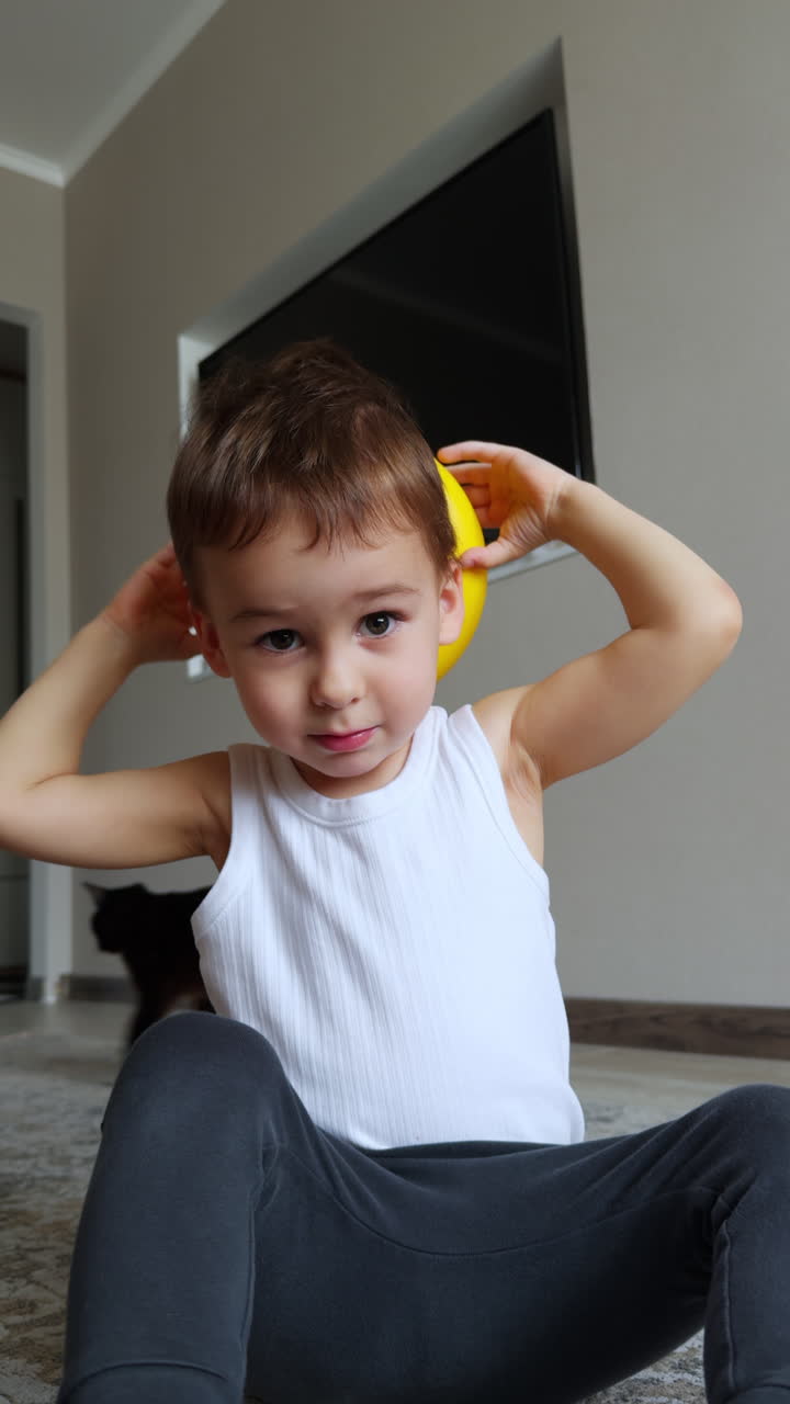 Adorable toddler sits on the floor. Kid places the toy on his head. Black cat walks around the child. Vertical video.