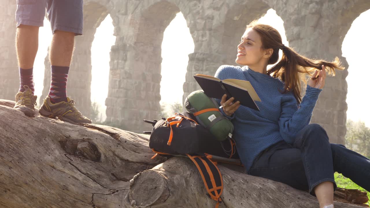feliz mujer joven mochilero turistas sentados tendidos en un tronco de tronco leyendo un libro frente a las antiguas ruinas del acueducto romano en el parque degli aquedotti en roma al amanecer pareja romántica en cámara lenta