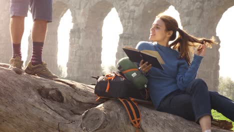 happy young woman backpacker tourists sitting lying on a log trunk reading book in front of ancient roman aqueduct ruins in parco degli acquedotti park in rome at sunrise romantic couple slow motion