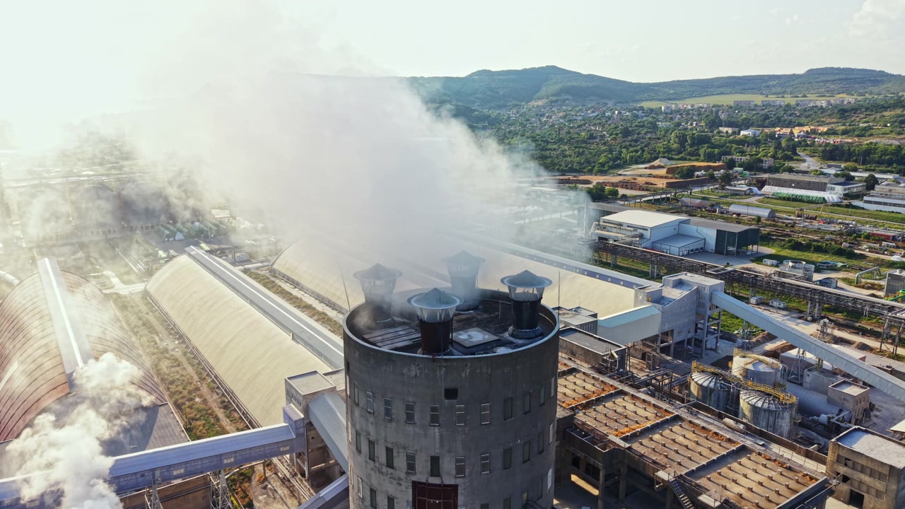 Fumo che sale da un edificio di fabbrica in una zona rurale di giorno, con colline visibili sullo sfondo e strutture che circondano l'edificio principale