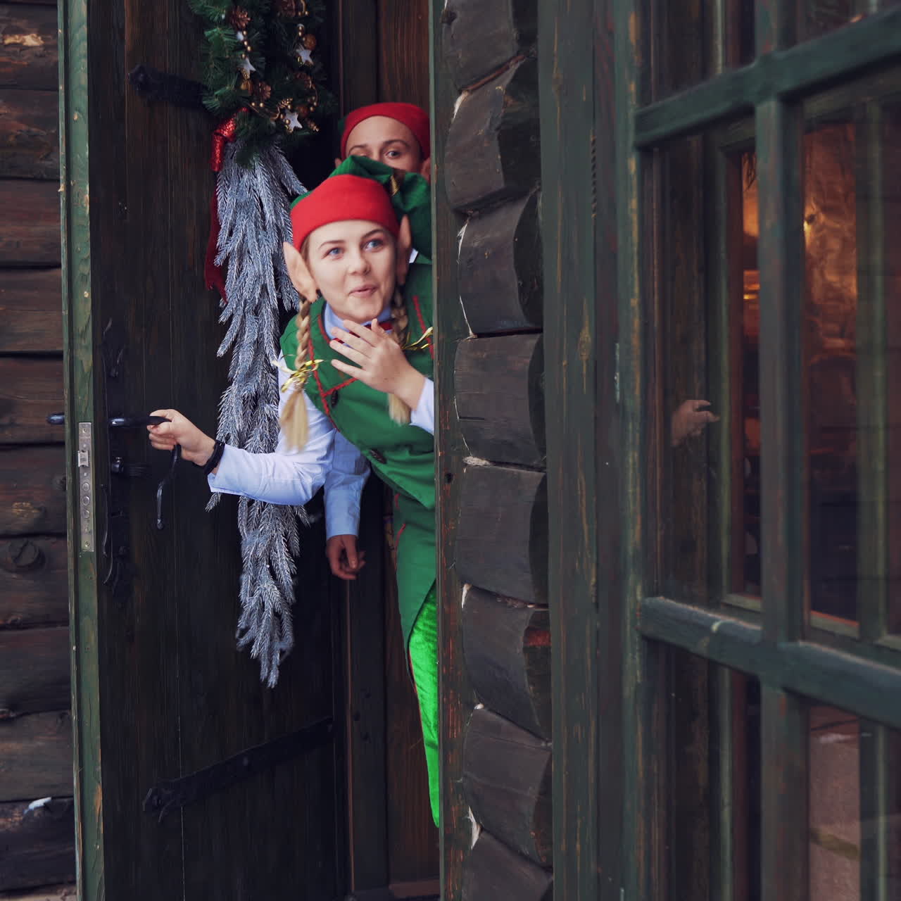 Happy elves peeking out from behind the door and laugh joyfully. Young female elf opens the front door inside the house.