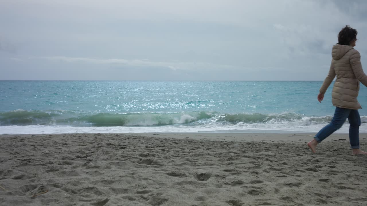 바다 파도가 모래 해변에 부 ⁇ 히고 겨울 재 ⁇ 을 입은 여자가  ⁇ 고 있습니다.