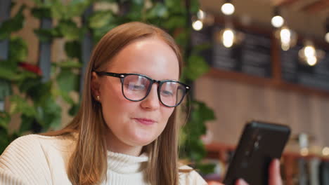 surprised and excited young woman in glasses smiles while chatting on smartphone in indoor cafe with leafy green decor, warm lighting, and blurred background featuring menu boards and glowing bulb