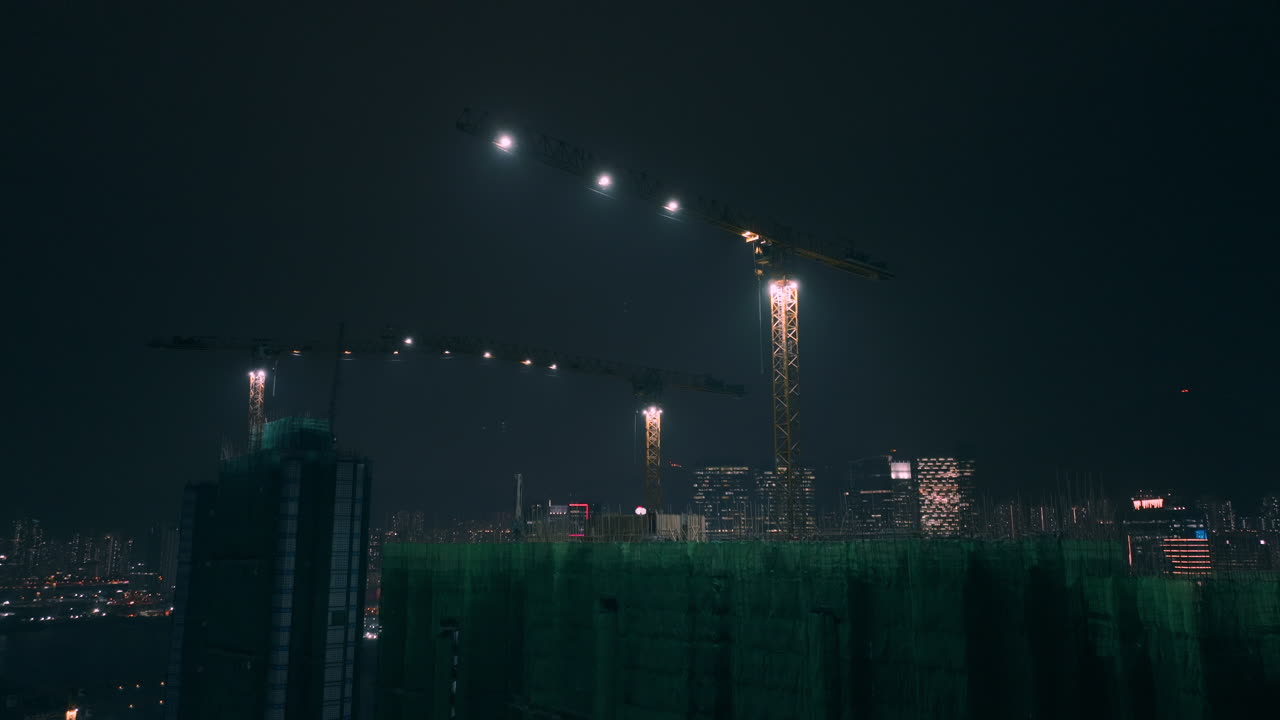 Aerial ascending shot showing cranes on top of residential building construction with Kowloon Bay district skyline in background at night - Hong Kong,Asia