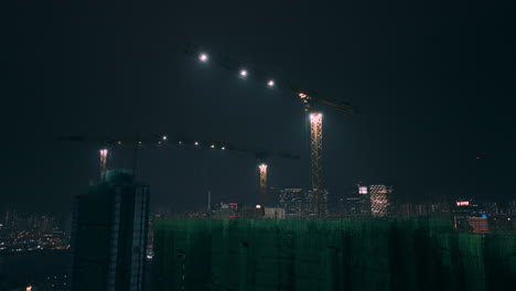 Aerial-ascending-shot-showing-cranes-on-top-of-residential-building-construction-with-Kowloon-Bay-district-skyline-in-background-at-night---Hong-Kong,Asia