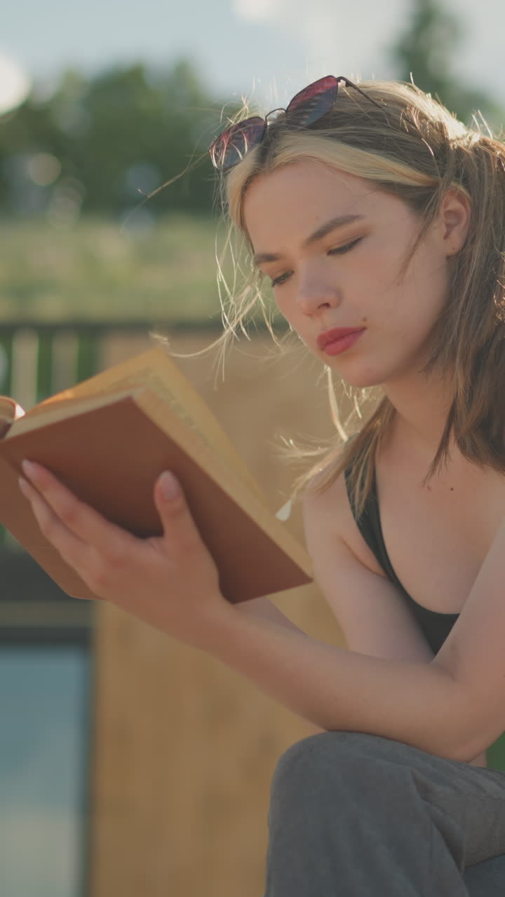 mujer sentada con las piernas cruzadas, leyendo un libro mientras el viento sopla a través de su cabello, su mano descansa en el libro, y una bolsa negra está a su lado, el fondo presenta una colina cubierta de hierba