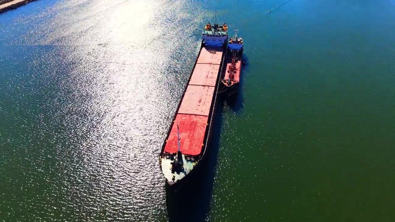 Buque granelero con una embarcación de apoyo en el mar. Toma en picado de un gran buque comercial con cubierta roja flotando en agua resplandeciente bajo la luz del sol.