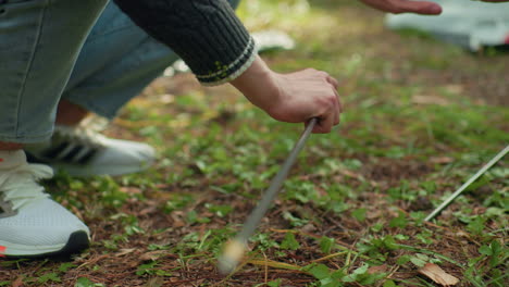 close up of man kneeling on forest ground assembling tent pole while friend hands over another pole, surrounded by green foliage and camping gear during calm outdoor setup moment