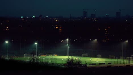 aerial cinematic drone view of a lit soccer field at night. football players are leaving the pitch after practice. city buildings in the background. top view.