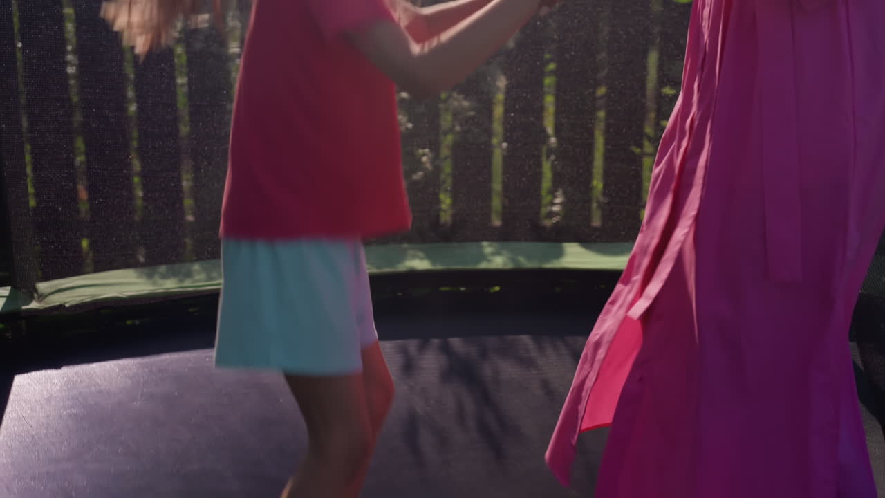 Young Girl Bounces Energetically On Trampoline, Child Enthusiastically Springs Through Sunlight Shadows In Backyard Play, Spirited Young Girl Leaps With Joy On Trampoline Near Her Mother Outside