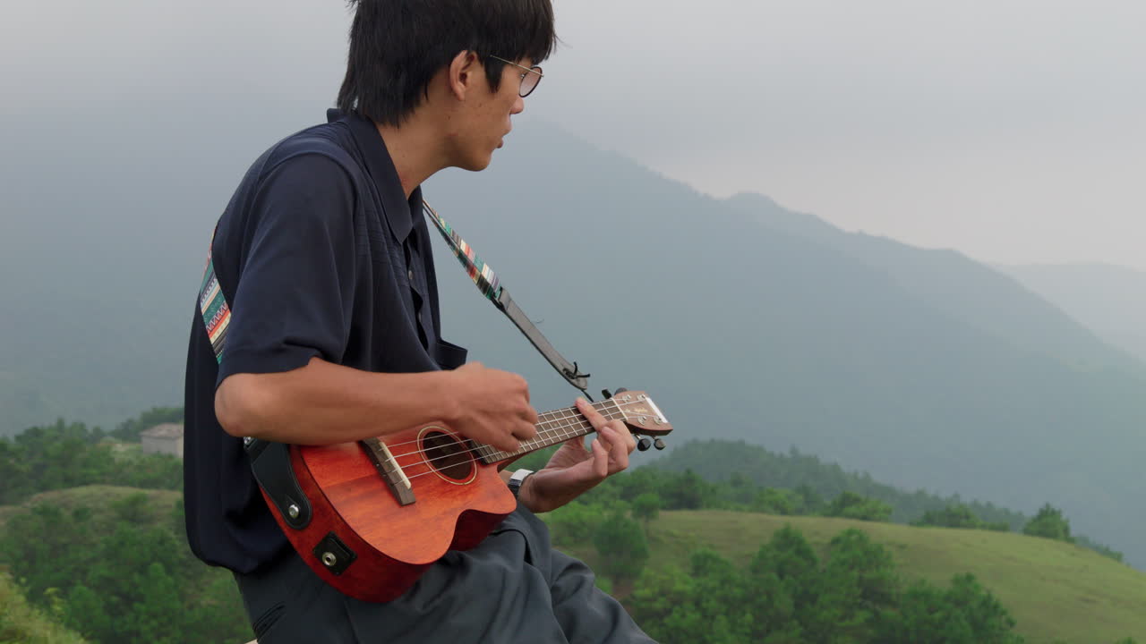 joven asiático elegante con gafas tocando el ukelele en la cima de una montaña tropical