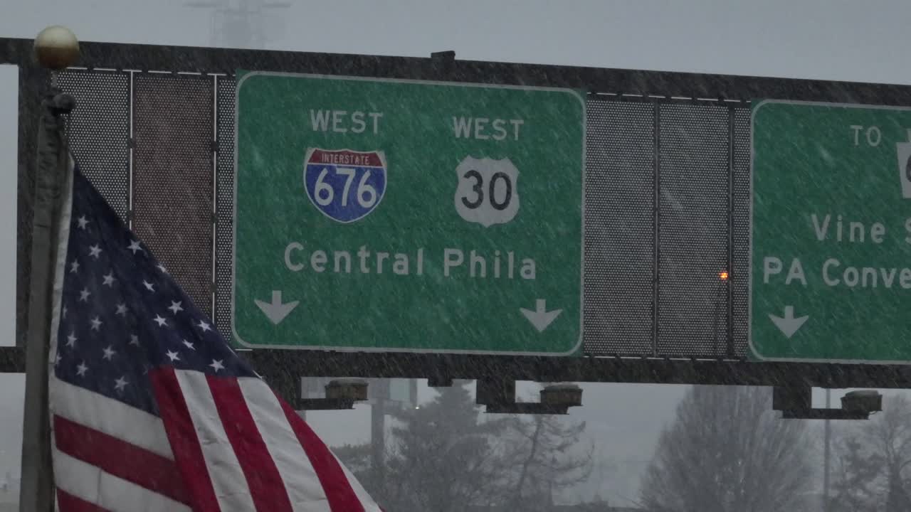 Amerikansk flag, der vajer under snefald ved siden af motorvejsskilte for Interstate 676 og Route 30, der fører til Central Philadelphia under vinterstormforhold. Luftfoto tæt på.