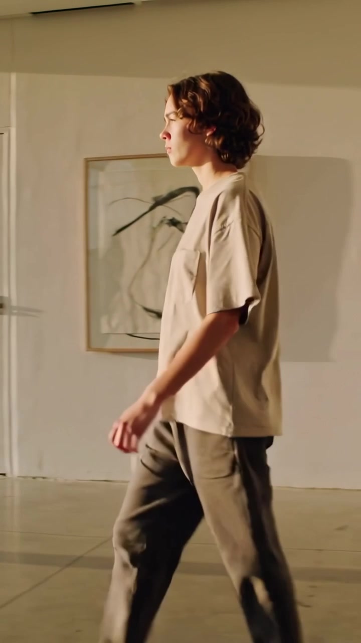Lodret video: Mand i T-shirt kommer ind fra venstre og går i studie, stopper ved vinduet og kigger på trætoppe.
