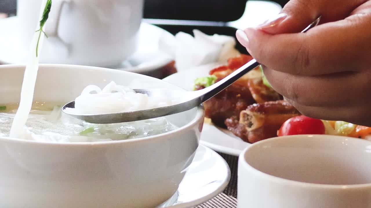 Close-up of hands using chopsticks and spoon to enjoy a noodle dish with fresh ingredients.