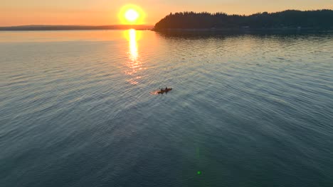 Vista-Aérea-De-Drones-Volando-En-El-Enfoque-De-Una-Pareja-Joven-Remando-Hacia-La-Puesta-De-Sol-En-Un-Kayak-De-Mar-En-La-Bahía-Cerca-De-Seattle,-Washington-Al-Amanecer