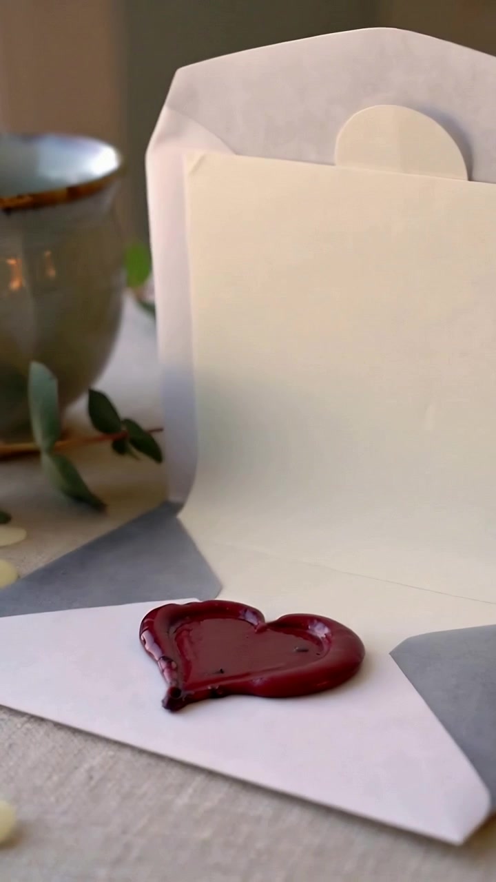 Vertical video: Lifting flap, hand revealing glossy red wax heart on white envelope on tabletop